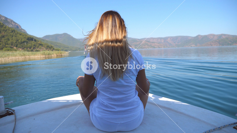 Rear back view of young girl sitting on bow of boat and looking to beautiful nature landscape on sunny day. Happy woman with blonde hair enjoying summer travel. Vacation or holiday concept. Close up.