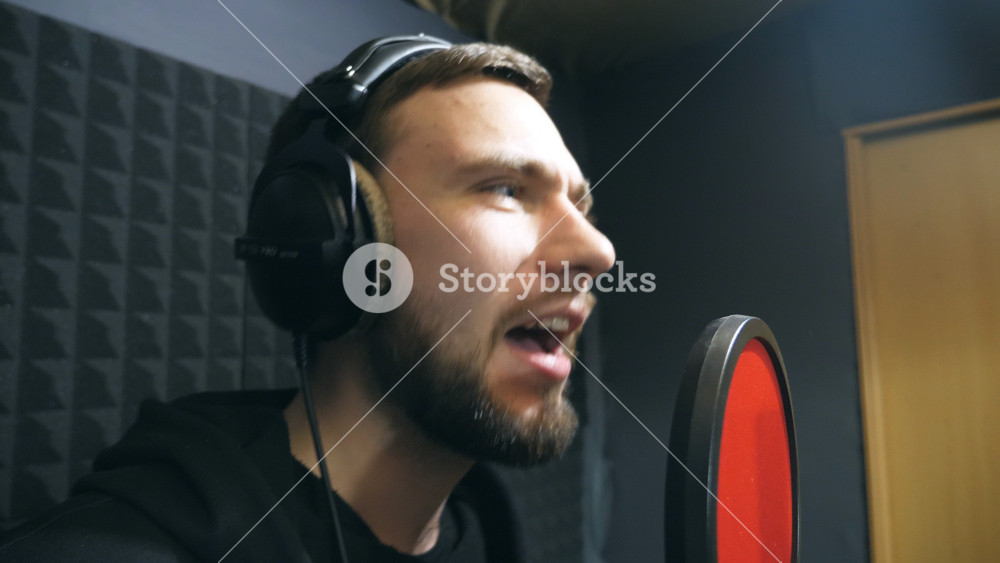 Male singer in headphones singing song into the microphone at sound studio. Young man emotionally recording new melody or song. Working of creative musician. Show business concept. Slow motion.