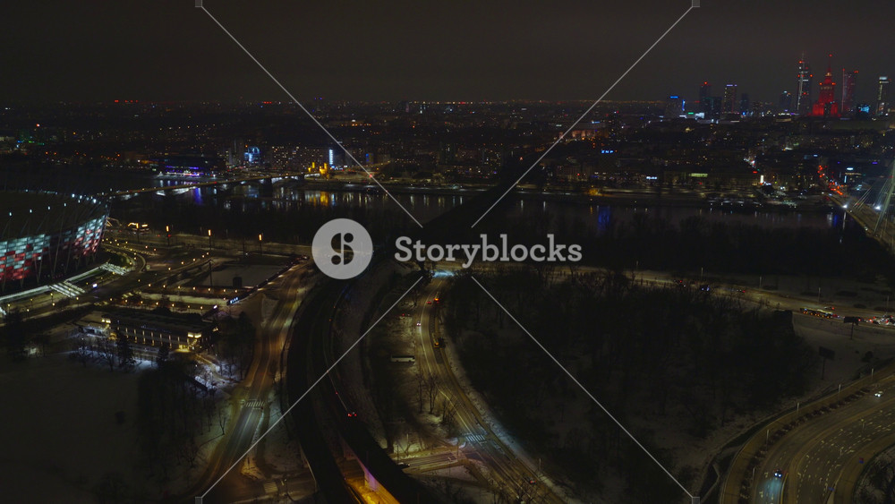 Aerial night over Warsaw, Poland shows Vistula River, PGE Narodowy, Swietokrzyski Bridge, snow dusted streets, amber lit station, moving cars and a passing train.