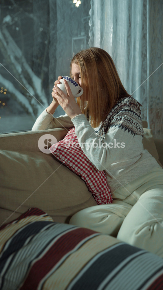 A cozy winter. A young beautiful woman admiring the festive lights outside the window, sitting with a cup of hot coffee tea on Christmas Eve