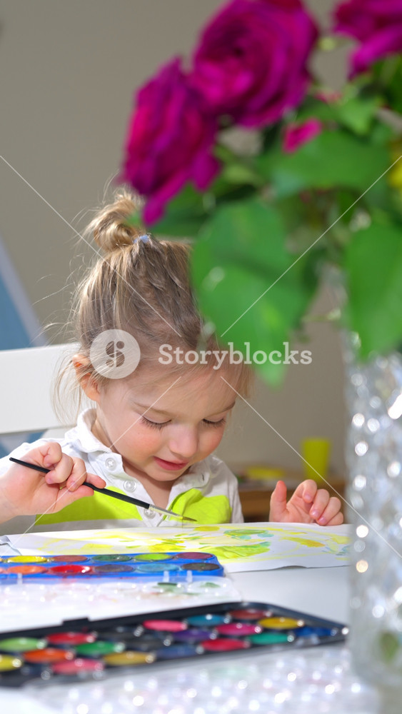 A young child and a woman painting together at a table with watercolor paints. The child smiles while holding a brush, and the woman assists. Cozy home setting with toys, plants, and decorations.