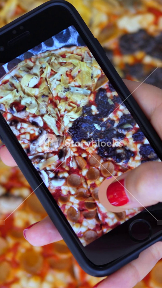 Close up of a female hand holding a smartphone and recording a video of a freshly baked pizza with different toppings, a concept for food