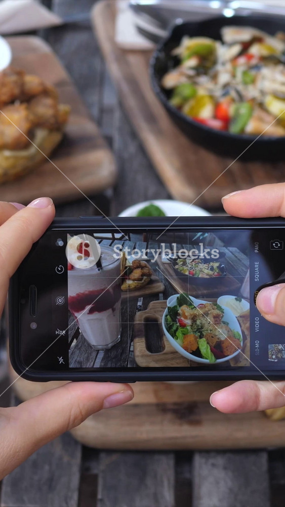 Close up of a food blogger's hands holding a smartphone, capturing images of a delicious lunch spread on a rustic wooden table for social