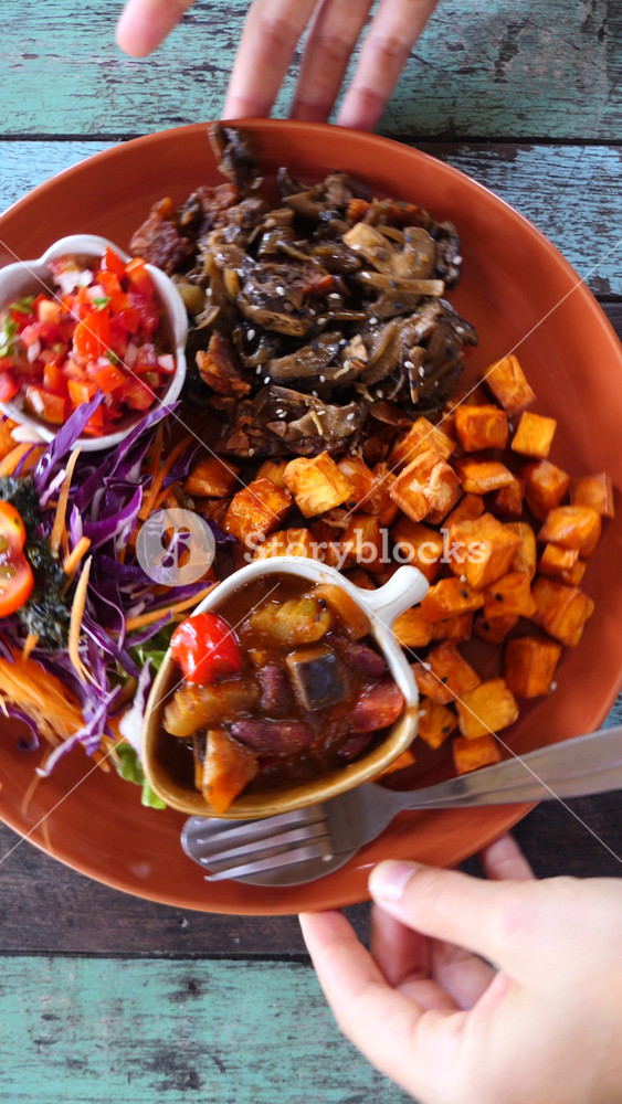 Colorful plate of plant based food including sweet potatoes, mushrooms, and fresh salad being placed on a rustic wooden table by a person